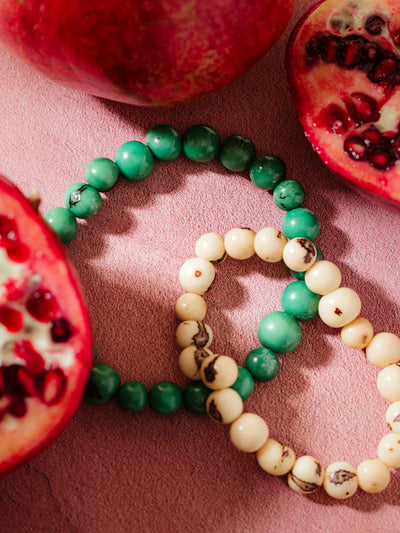 Two handcrafted beaded bracelets, one featuring vibrant green stones and the other with cream-colored beads, styled with fresh pomegranate halves and a textured pink background, showcasing bold and natural artisan jewelry.