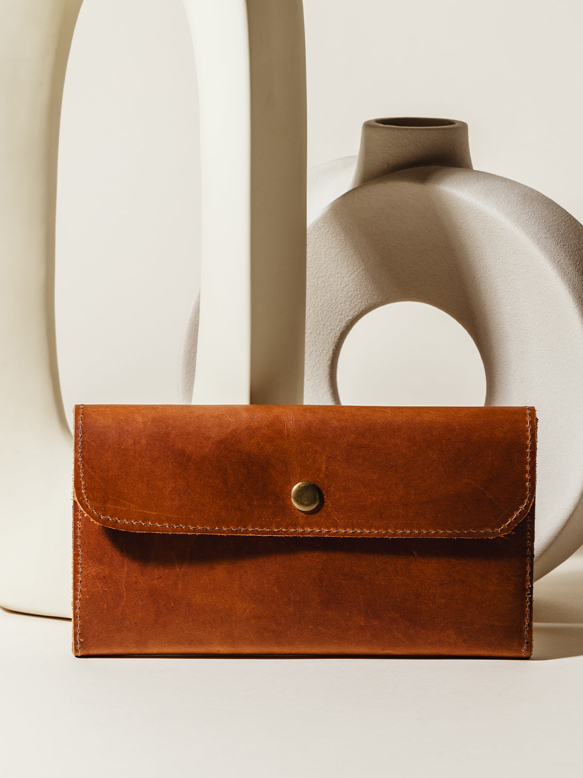 A classic brown leather wallet with a brass snap closure displayed against modern ceramic decor, highlighting its minimalist and durable design.