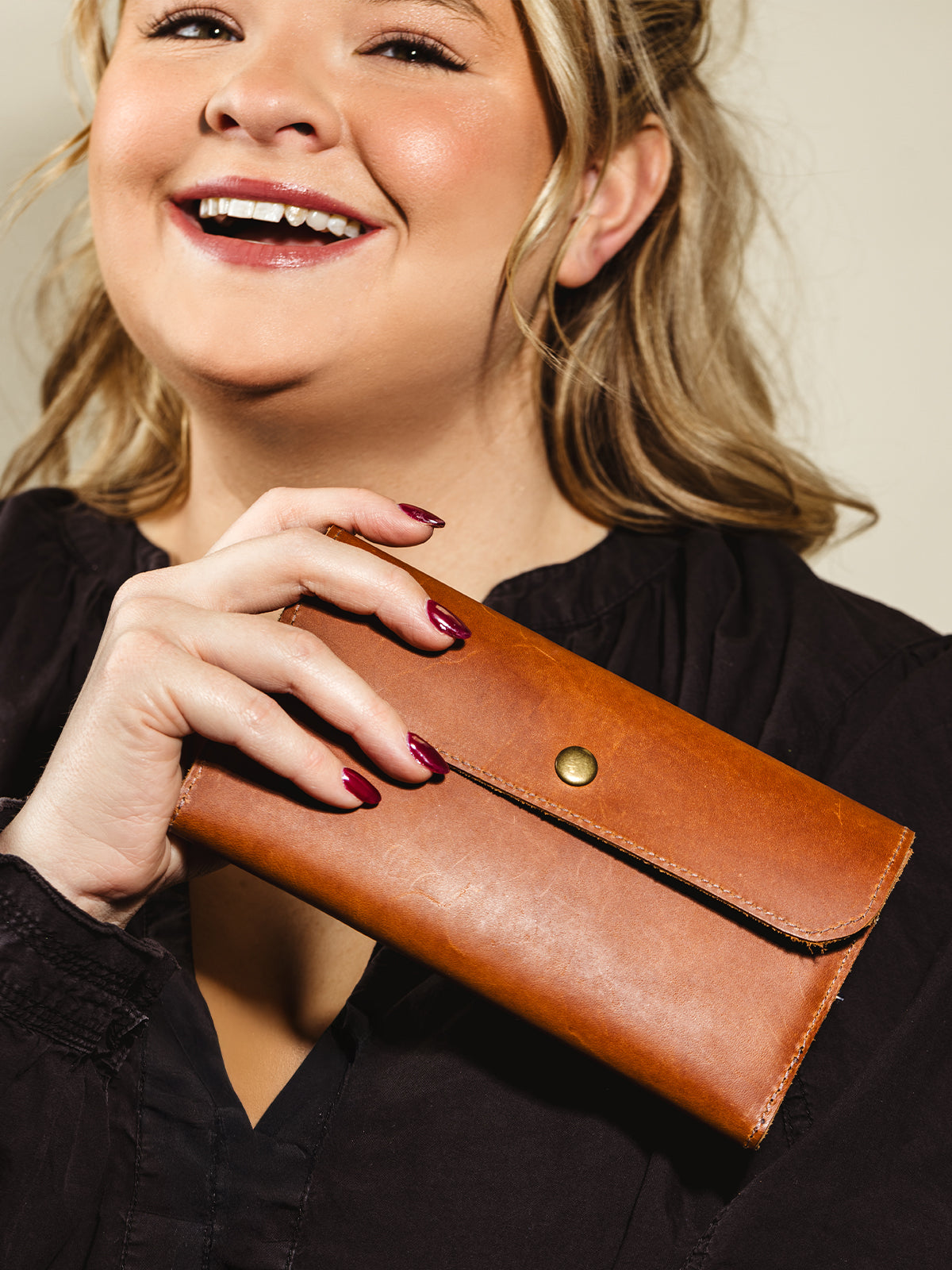 A smiling woman holding a brown leather wallet with a brass snap closure, styled with a black outfit for a timeless and versatile accessory look.