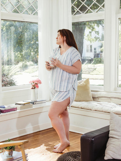 Woman holding a mug, gazing outside while wearing Swahlee's striped pajama shorts and top. Ethically crafted sleepwear perfect for cozy mornings.