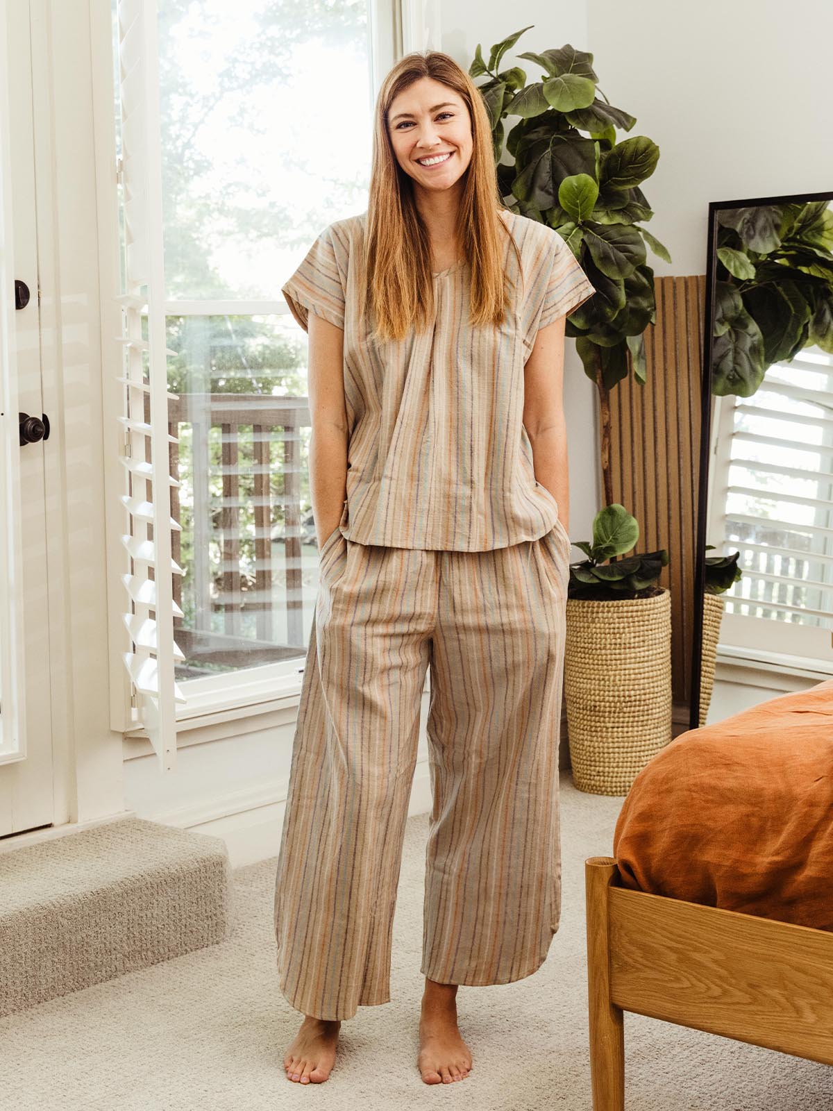 Woman standing barefoot in a bedroom, wearing wide-leg striped pajama pants and a matching top from Swahlee. Sustainable and ethical sleepwear for women.