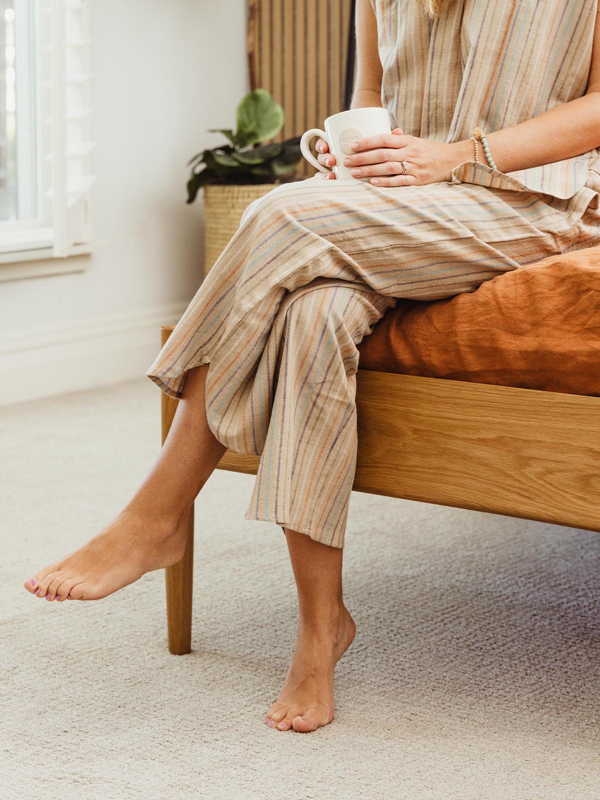 Woman sitting on the edge of a bed, holding a mug, wearing Swahlee's striped loungewear set. Sustainable pajamas for slow, relaxed mornings.