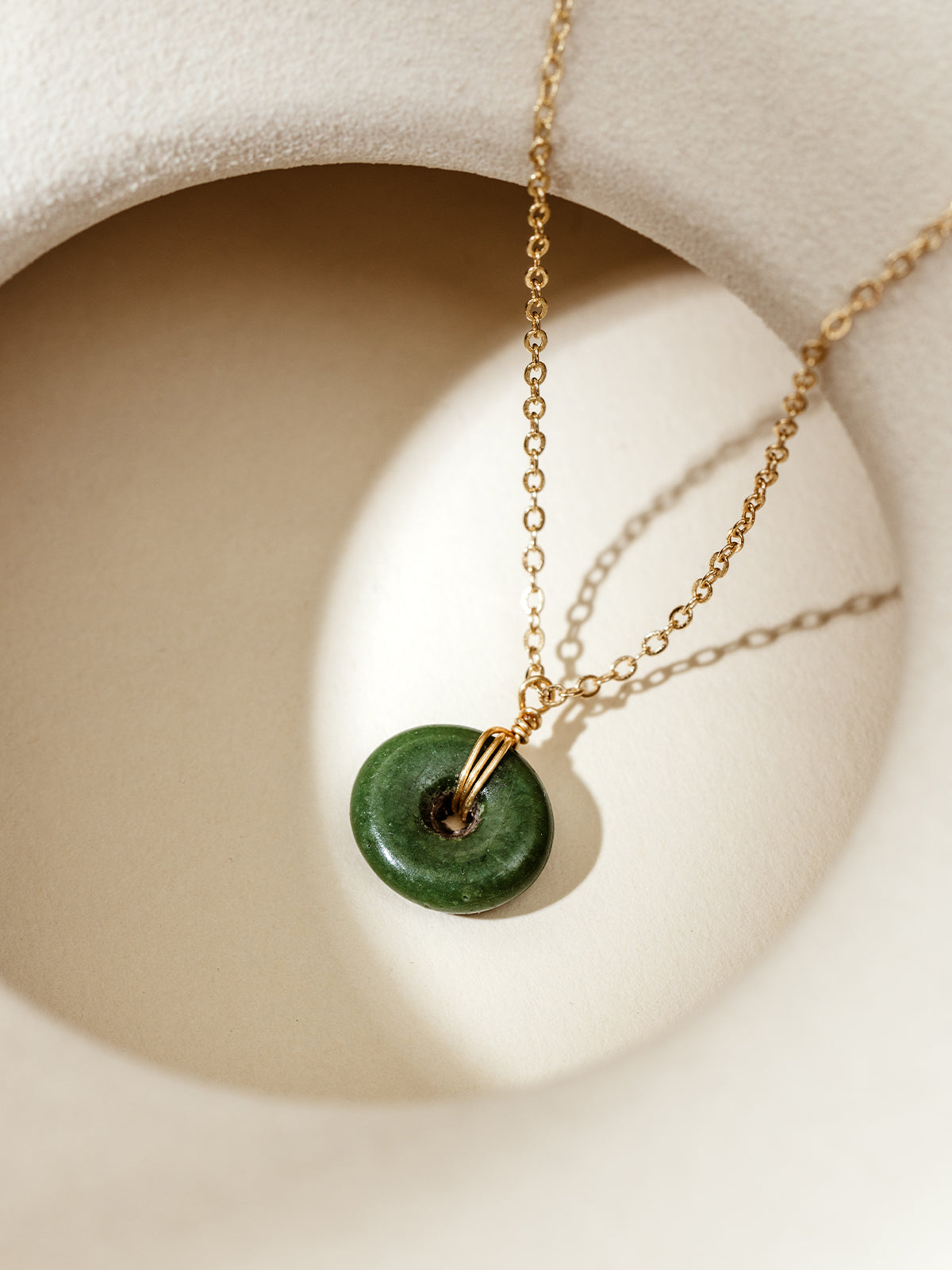 Minimalist gold chain necklace with a smooth green glass donut pendant, styled on a textured white surface with soft natural lighting, emphasizing artisan craftsmanship and sustainable elegance.