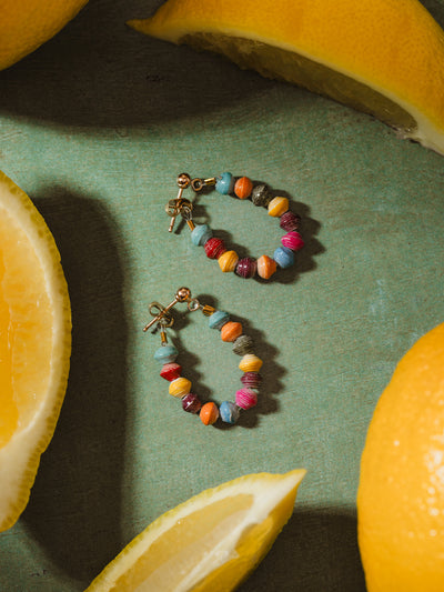 A pair of colorful beaded hoop earrings with gold accents, featuring vibrant handcrafted beads in shades of blue, red, yellow, and orange, artfully arranged on a green background with fresh lemon slices, creating a fresh and lively aesthetic.