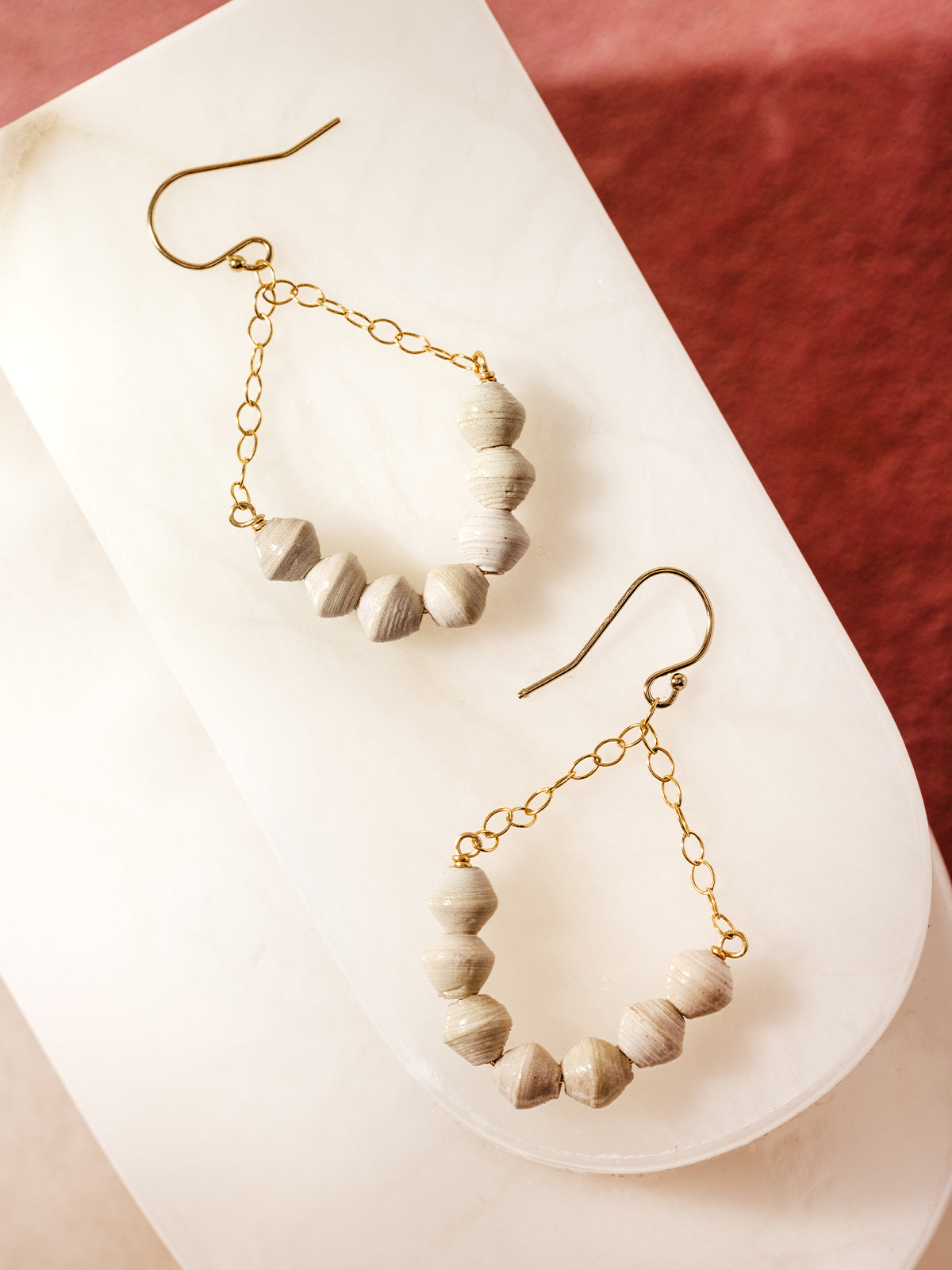 Handcrafted gold hook earrings featuring light gray recycled paper beads and delicate gold chain accents, styled on a white marble slab with a blush pink background, showcasing sustainable and elegant artisan jewelry.