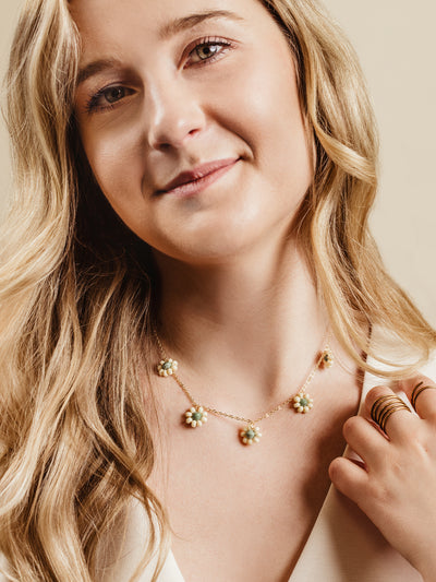 Close-up of a woman wearing a gold chain necklace adorned with delicate floral charms featuring white and green beads, styled with gold stackable rings and a cream blouse, highlighting handcrafted and elegant artisan jewelry.