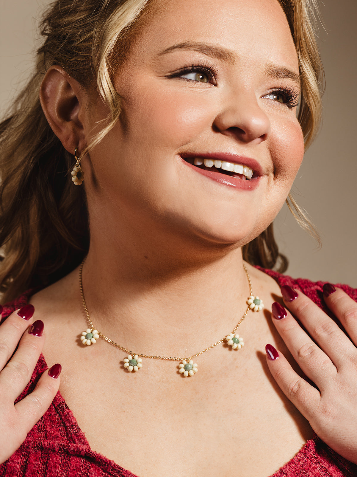 Smiling woman wearing a gold chain necklace with daisy-shaped charms and matching earrings, styled with a cozy red sweater, highlighting delicate and feminine handcrafted jewelry.