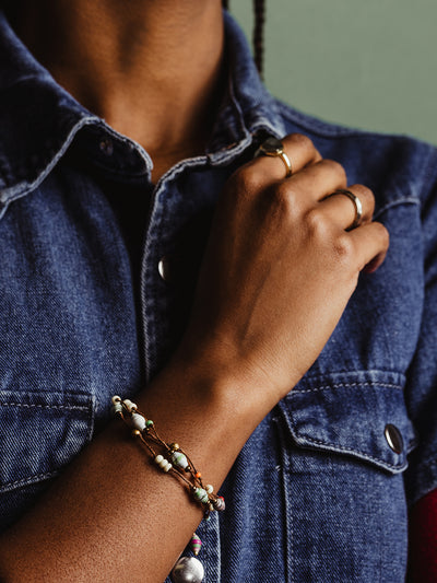 Close-up of a woman's hand wearing a handmade beaded bracelet featuring earthy tones and natural beads, styled with a denim jacket and gold rings, highlighting sustainable and ethical fashion accessories.