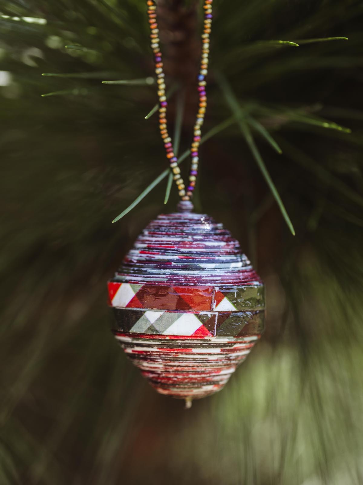 Handmade beaded ornament with colorful patterned strips, hanging from a multicolored beaded string on a green pine tree, adding a festive touch.