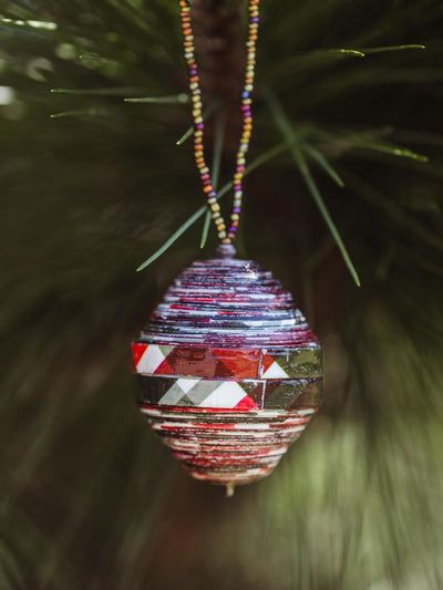 Handmade beaded ornament with colorful patterned strips, hanging from a multicolored beaded string on a green pine tree, adding a festive touch.