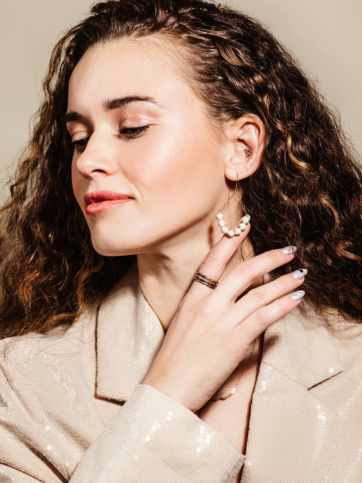 Close-up of a woman with curly brown hair wearing a beige sequined blazer, showcasing elegant handcrafted gold earrings with white beaded accents, a delicate gold necklace, and a stackable ring, highlighting a sophisticated and timeless jewelry look.