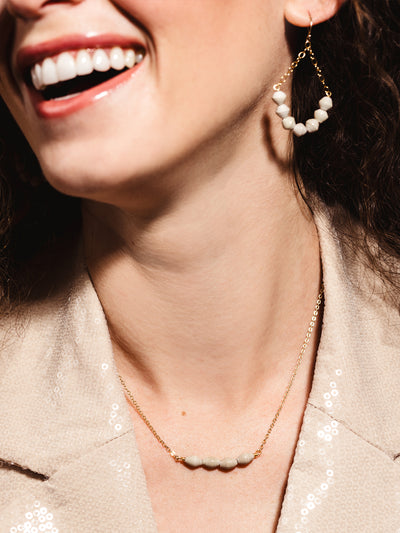 Close-up of a smiling woman wearing a gold chain necklace with five neutral-toned handmade beads and matching gold drop earrings featuring a beaded teardrop design, styled with a beige sequined blazer for an elegant and modern look.