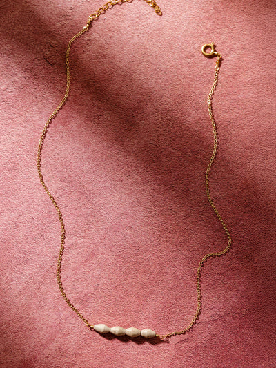 A delicate gold chain necklace featuring five handcrafted neutral-toned beads, elegantly laid on a soft pink fabric background, highlighting its minimalist and artisanal design.