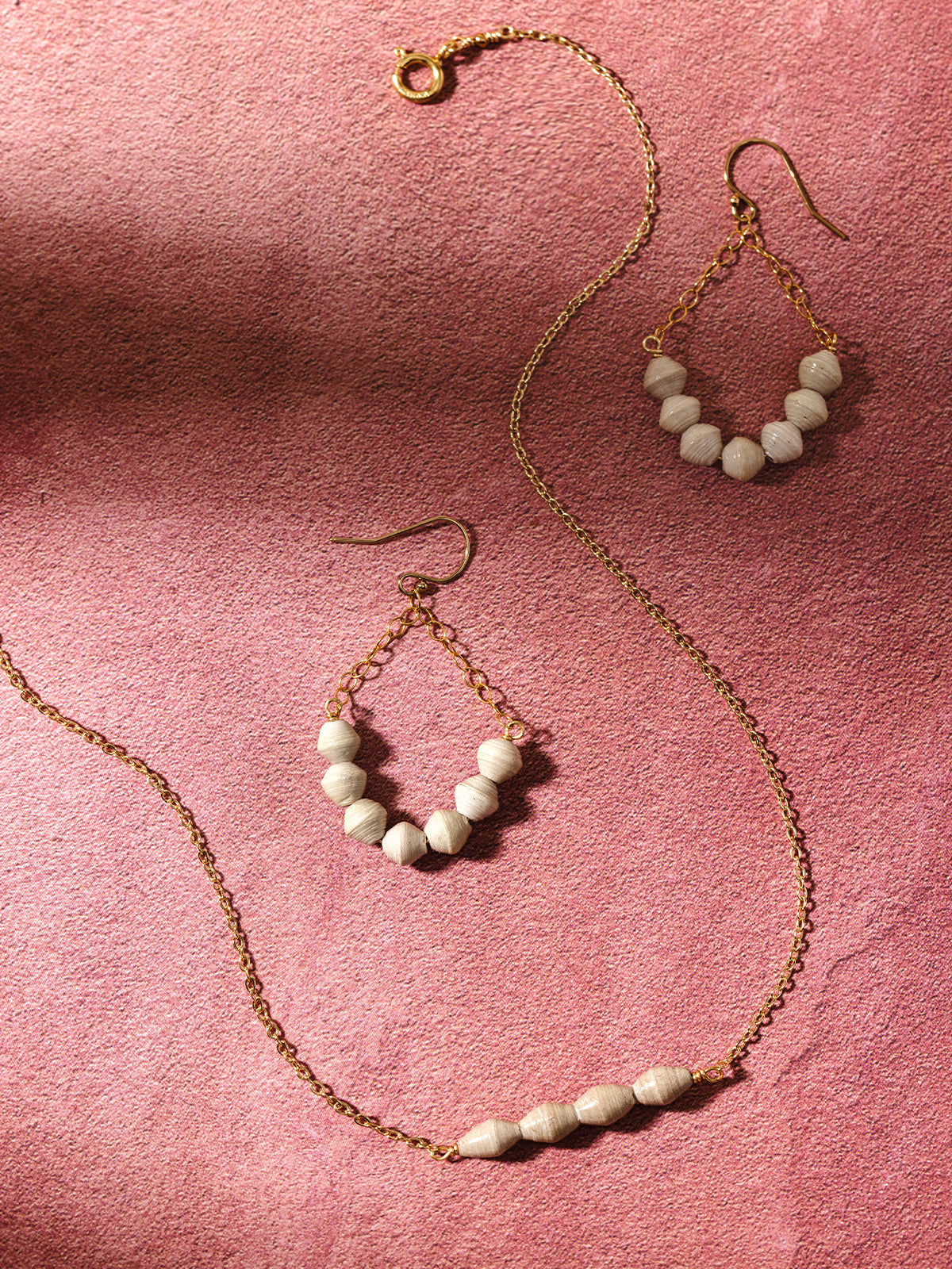 A gold beaded jewelry set, including a necklace and matching teardrop earrings with neutral-toned handcrafted beads, displayed on a pink textured background, showcasing their timeless and elegant design.