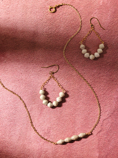 A gold beaded jewelry set, including a necklace and matching teardrop earrings with neutral-toned handcrafted beads, displayed on a pink textured background, showcasing their timeless and elegant design.