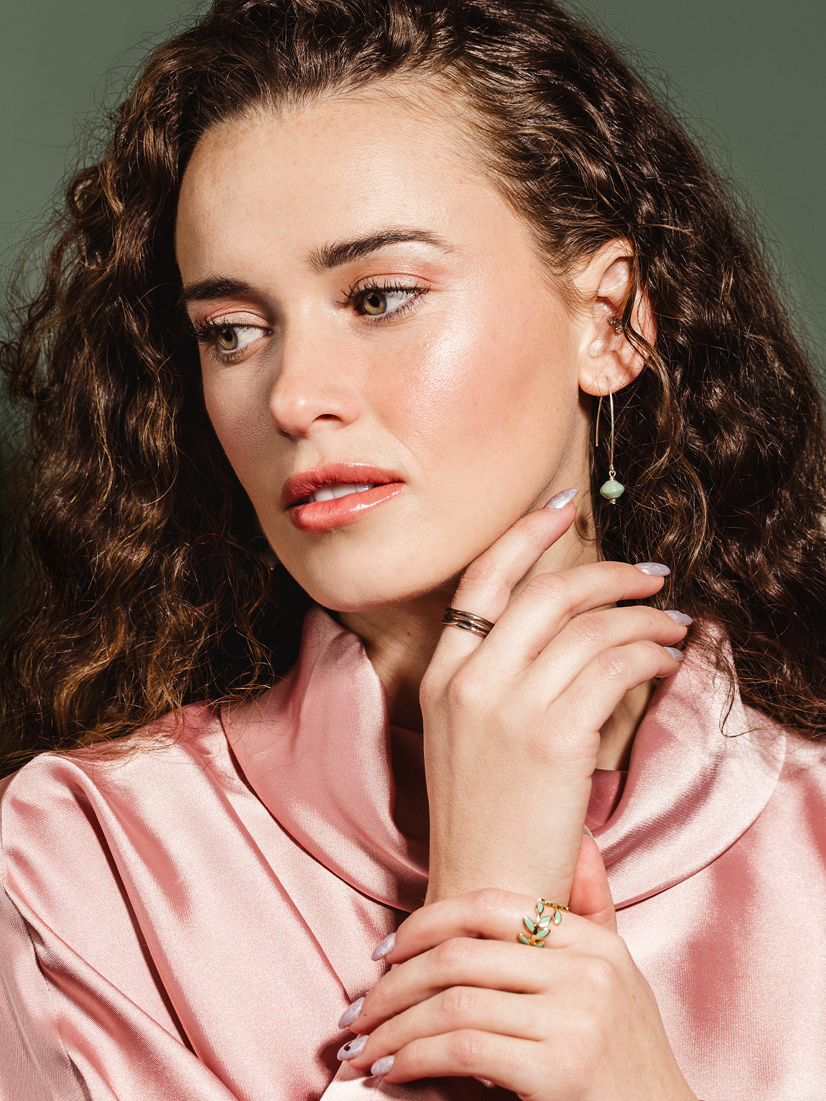 A woman wearing a pink satin blouse, accessorized with silver threader earrings featuring green handcrafted beads and a gold leaf ring with enamel accents, showcasing a chic and sophisticated style.