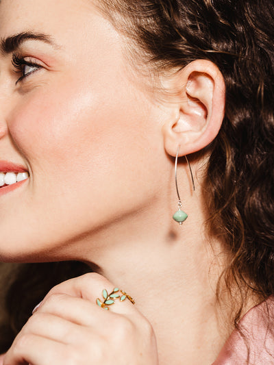 A close-up of a smiling woman wearing a silver threader earring with a light green handcrafted bead, paired with a gold leaf ring featuring green enamel details, adding a touch of elegance to her look.