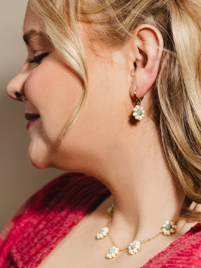 Close-up of a woman with blonde hair wearing a floral-themed jewelry set, featuring gold hoop earrings and a matching necklace adorned with handcrafted cream and green beaded flowers, styled with a cozy pink knit sweater for a charming, feminine look.