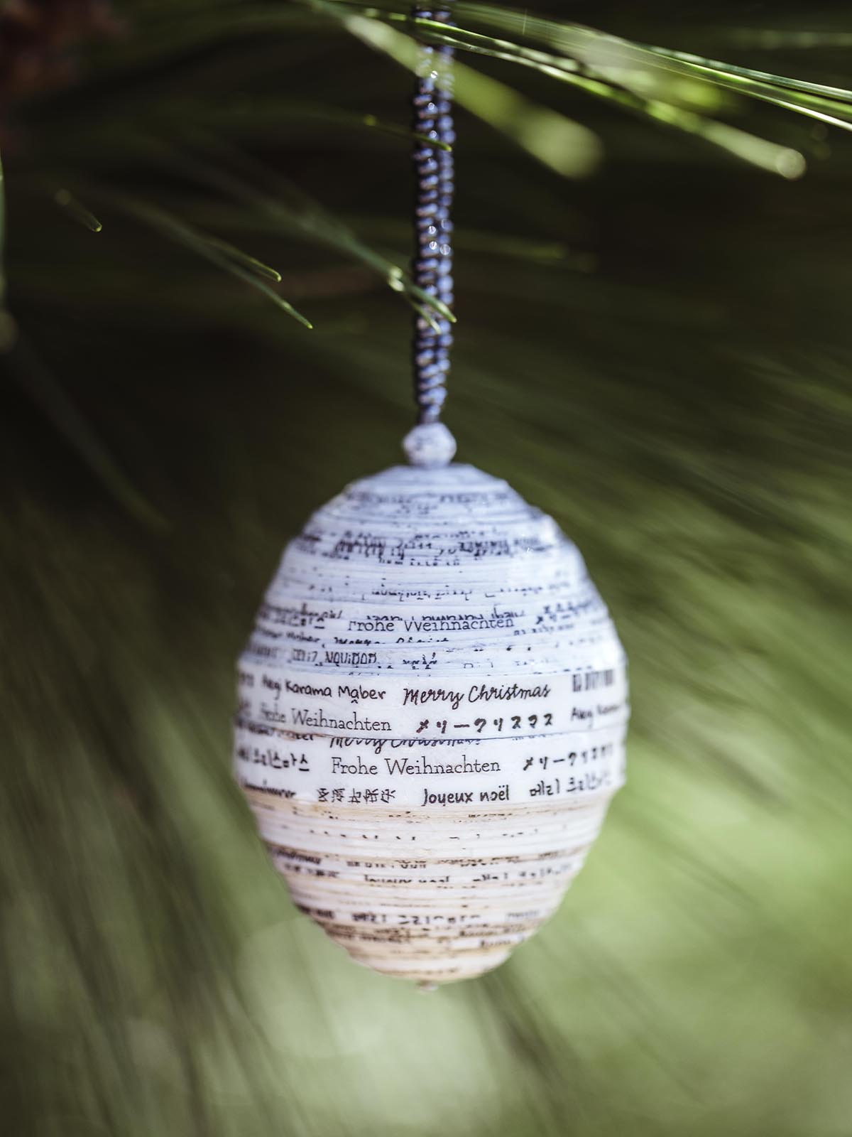 Handcrafted paper ornament featuring 'Merry Christmas' written in multiple languages, hanging from a dark beaded string on a green pine tree.