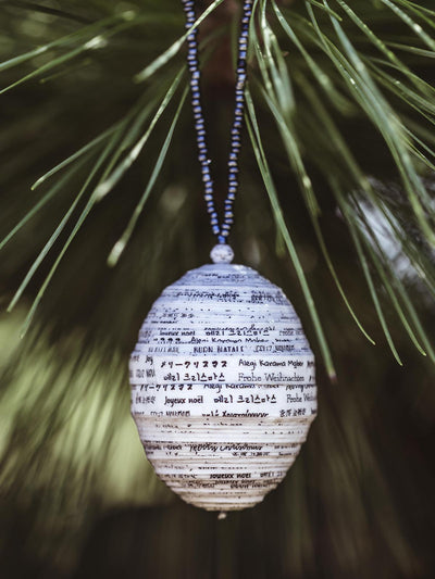 White handcrafted paper ornament with multilingual 'Merry Christmas' greetings, hanging from a dark beaded string on a pine tree branch.