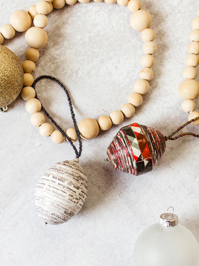 Handcrafted Christmas ornaments made from upcycled materials, featuring a white paper-bead ornament with printed text and a red and silver beaded ornament, displayed alongside wooden garland and a frosted glass bauble, part of Joffa.com's ethically sourced holiday decor collection.