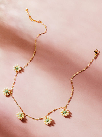 Delicate gold chain necklace adorned with dainty floral charms featuring white bead petals and soft green centers, styled on a blush pink backdrop with natural lighting, highlighting artisan craftsmanship and sustainable elegance.