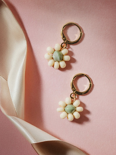 Handcrafted floral earrings with white bead petals and soft green centers, set on gold hoops, styled against a blush pink textured background with a satin ribbon accent, showcasing artisan and feminine design.