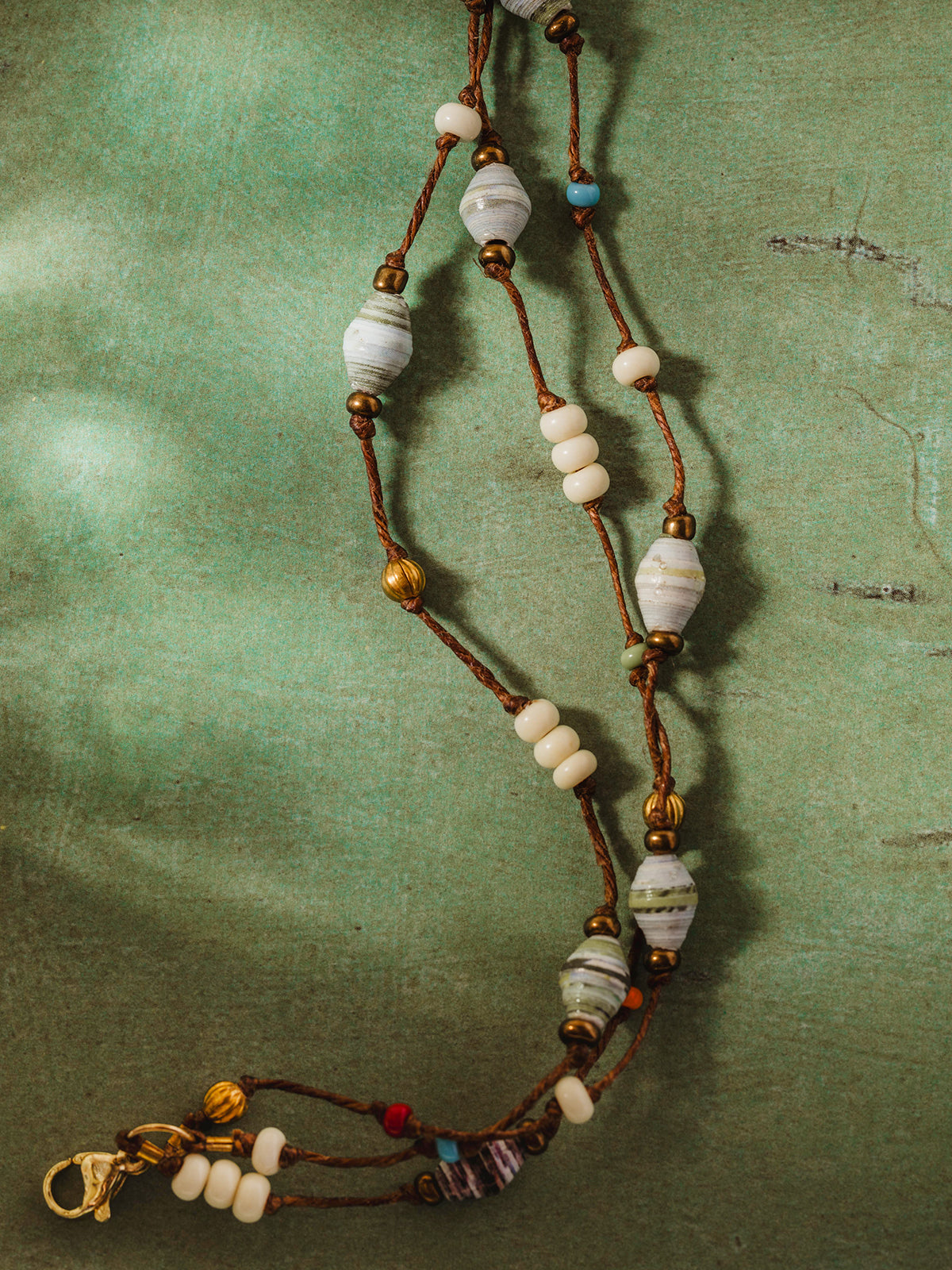 Handcrafted necklace featuring recycled paper beads in soft pastel hues and white accents, arranged on a green textured background with warm natural lighting, showcasing eco-friendly and artisan jewelry design.