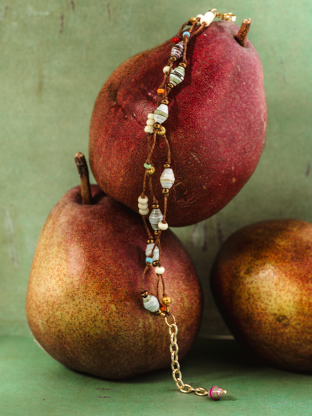 Handmade recycled paper bead necklace styled on deep red and golden pears, set against a muted green backdrop, highlighting sustainable and elegant artisan craftsmanship.