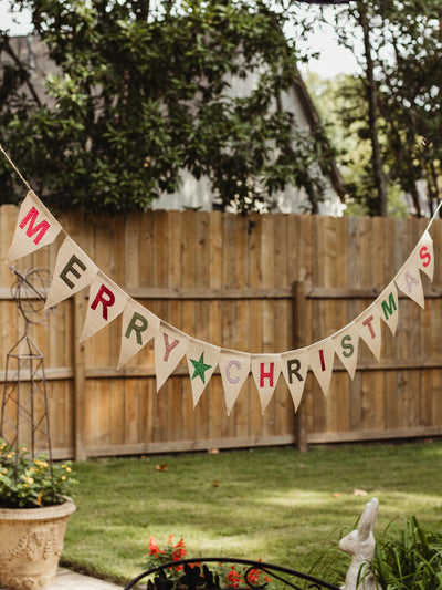 Burlap 'Merry Christmas' banner hanging across a garden space with a wooden fence in the background, set in a warm outdoor setting.