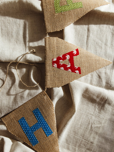 A detailed close-up of a rustic burlap pennant banner featuring hand-stitched fabric letters, laid out on a soft beige linen backdrop.