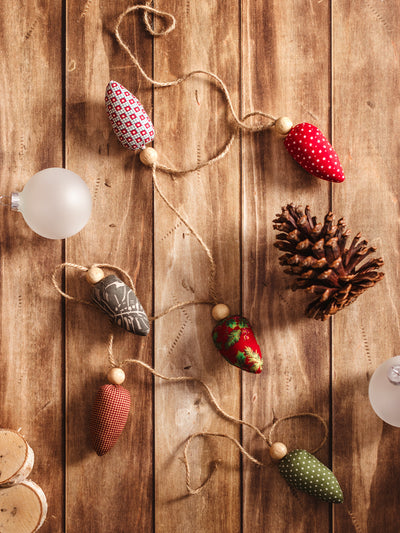 Handmade fabric Christmas lightbulb garland featuring colorful lightbulb-shaped fabric pieces and wooden beads, draped on a rustic wooden surface, adorned with a pinecone and frosted glass.