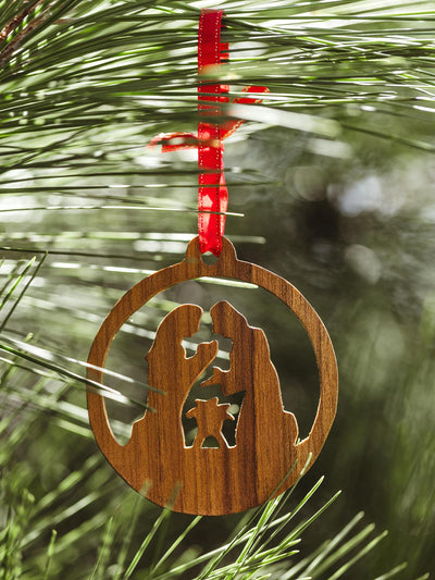 Handcrafted wooden nativity ornament featuring Mary, Joseph, and baby Jesus, hanging from a red ribbon on a pine tree branch, symbolizing the Christmas story.