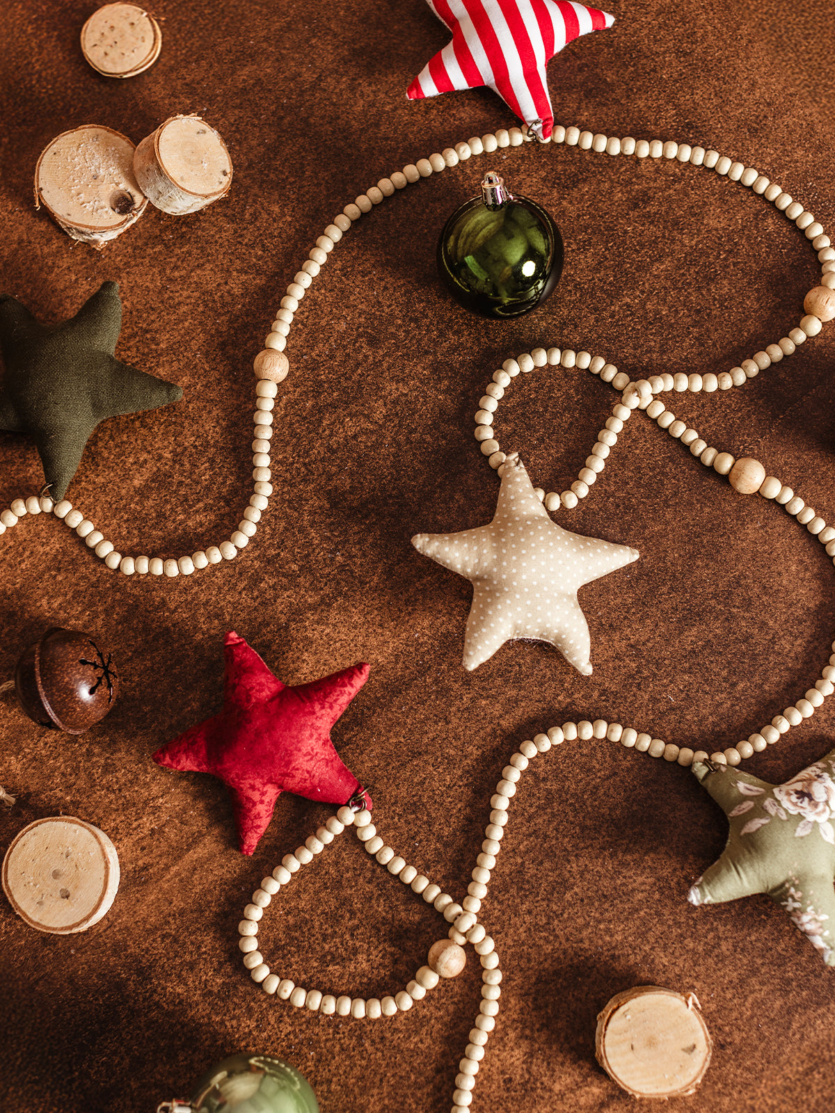 Handcrafted star-shaped fabric ornament garland with wooden beads and Christmas baubles, arranged on a wooden surface with small tree slices, creating a festive holiday display.