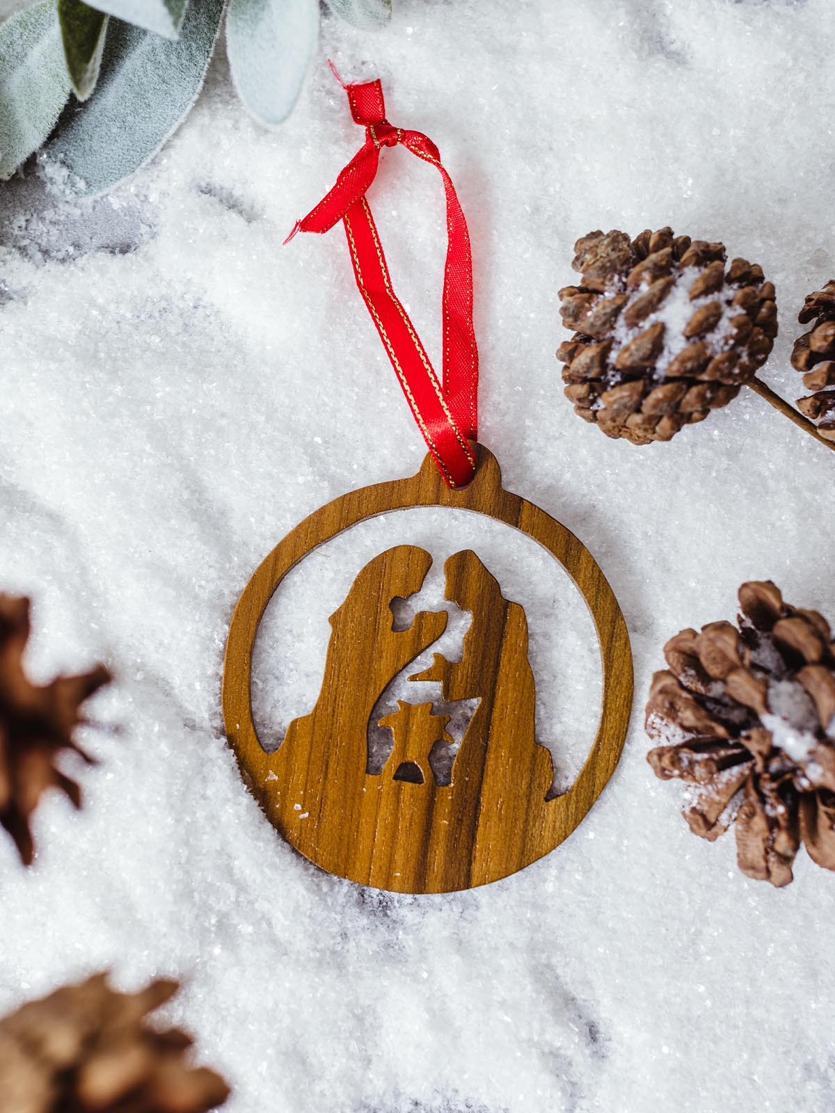 Handmade wooden nativity ornament with Mary, Joseph, and baby Jesus, placed on faux snow with pinecones and greenery, tied with a red ribbon for a rustic holiday display.