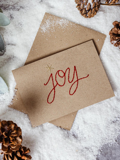 Sustainable holiday card with the word 'joy' embroidered in red on a kraft paper card, placed in faux snow alongside pinecones, perfect for a natural Christmas theme.