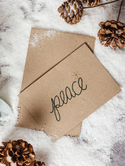 Eco-friendly holiday card with the word 'peace' embroidered in green on a kraft paper card, surrounded by pinecones and faux snow for a rustic, festive look.