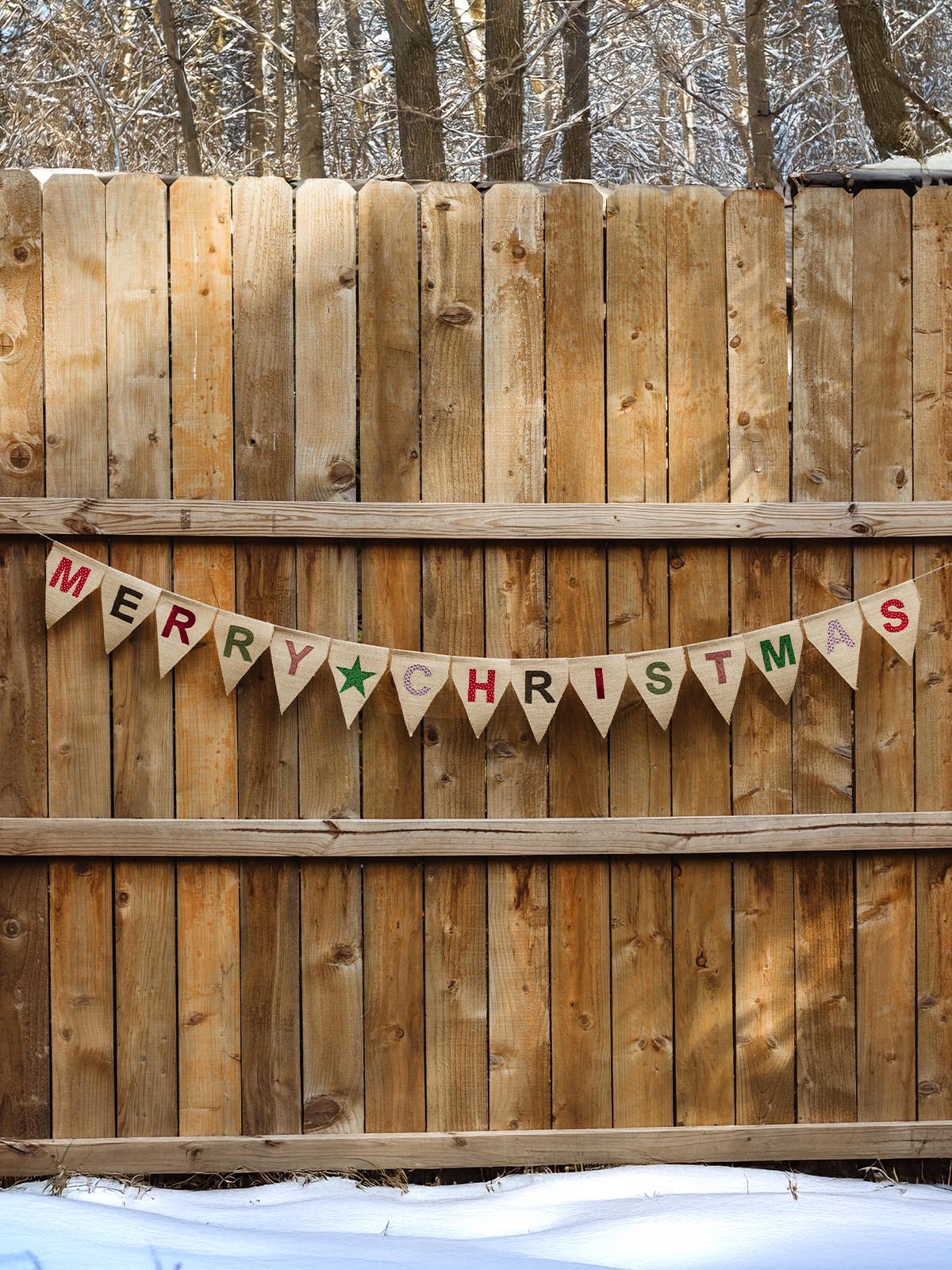 Handcrafted burlap 'Merry Christmas' banner hanging on a wooden fence, with a snowy outdoor background.