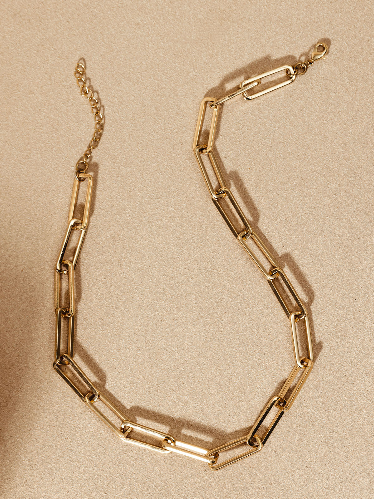 Gold chain necklace laid flat on a light tan surface, showcasing its rectangular links and simple clasp.