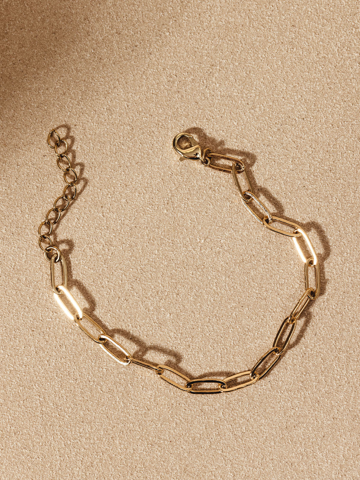 Gold chain bracelet with rectangular links laid flat on a light tan surface, showing its delicate clasp and extender chain.