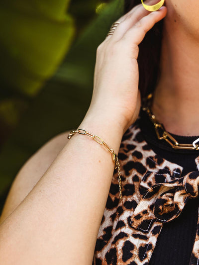 Close-up of a woman's wrist wearing a gold chain bracelet with rectangular links, paired with a leopard print top, as she gently touches her earring with her hand.