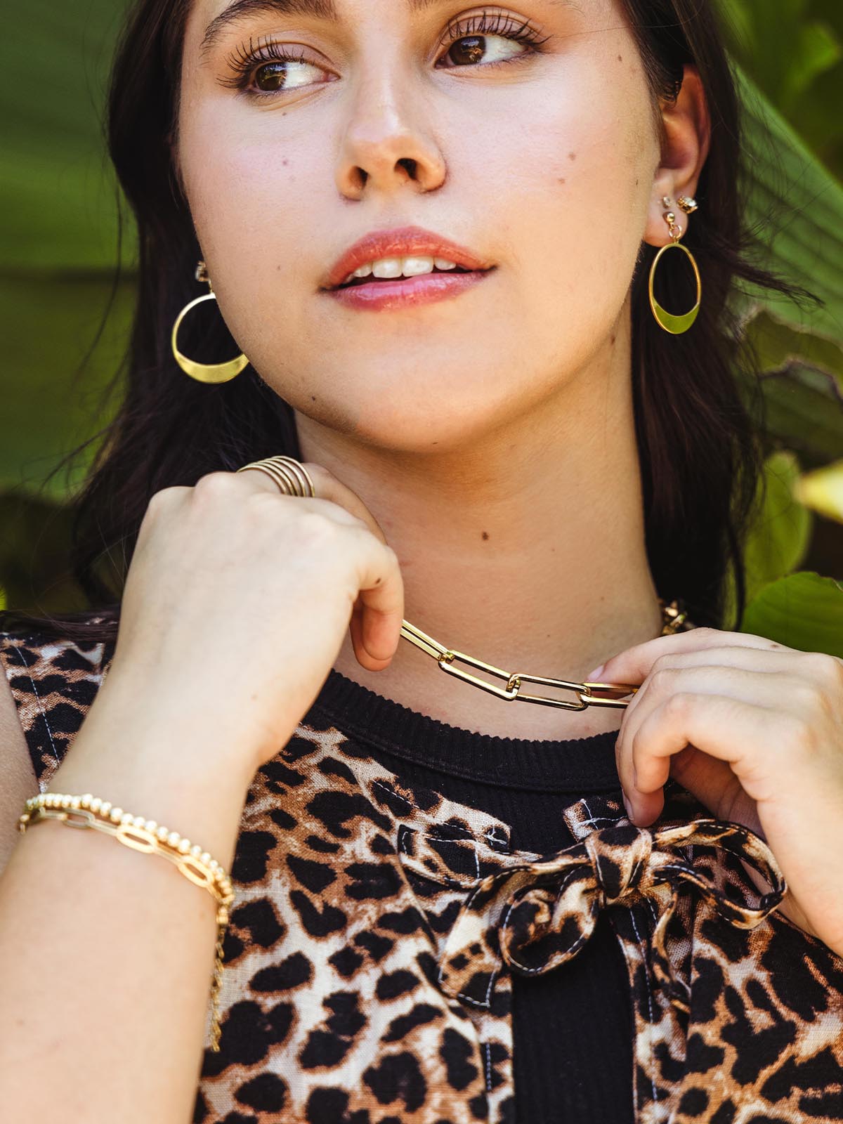 Close-up of a woman wearing a gold chain necklace with rectangular links, styled with a leopard print top, gold hoop earrings, and a gold bracelet, as she lightly touches the necklace.