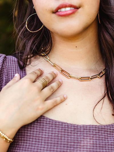 Close-up of a woman wearing a gold chain necklace with rectangular links, paired with a purple checkered top, a gold ring, and gold hoop earrings.