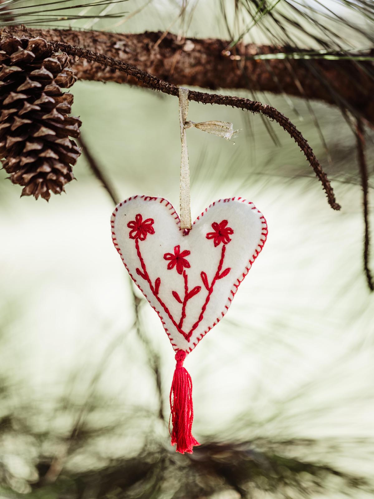 Handcrafted heart-shaped felt ornament with red embroidered flowers, hanging from a pine tree branch with a delicate ribbon, creating a festive and rustic holiday look.