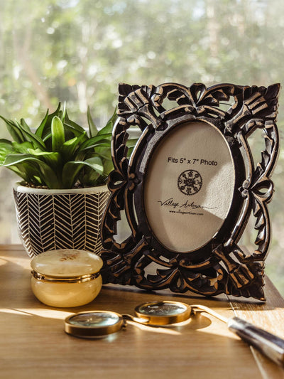 Village Artisan carved black wooden photo frame for 5x7 photos, displayed with a potted plant and gold accent items on a wooden surface. Ethical home decor available at Joffa.