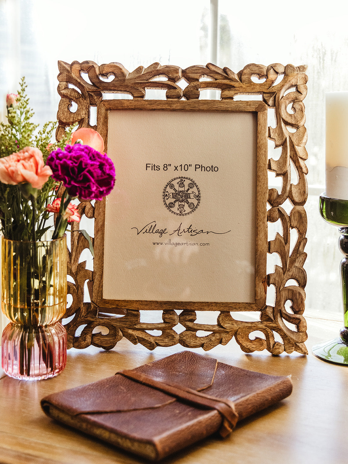 Village Artisan 8x10 carved wooden photo frame, styled with vibrant flowers and a leather-bound journal on a sunlit table. Fair trade home decor from Joffa.