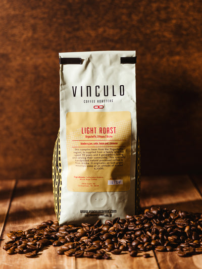 A Vinculo Coffee Roasters Light Roast coffee bag with a yellow label stands alone on a wooden surface, surrounded by roasted coffee beans. The label describes its origin from Yirgacheffe, Ethiopia, and its tasting notes.