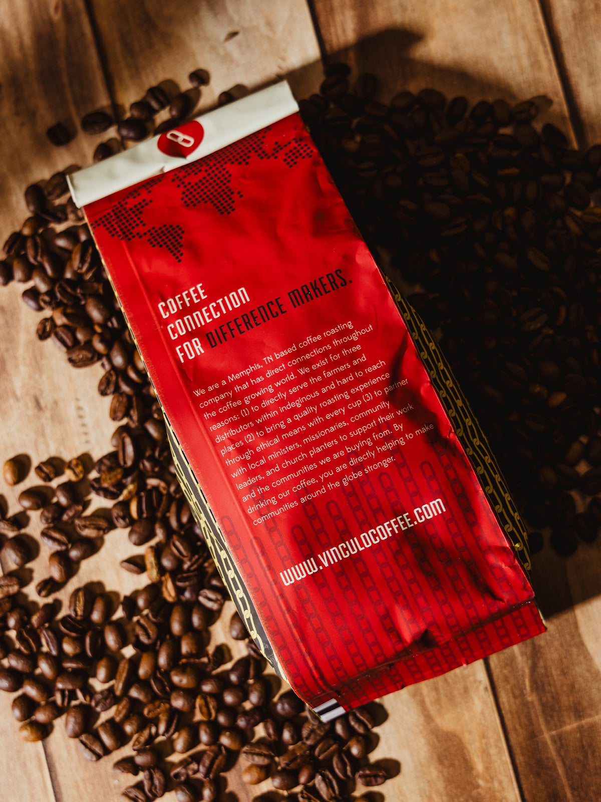 The back of a Vinculo Coffee Roasters bag featuring a bold red design with white text reading "Coffee Connection for Difference Makers." The description highlights the company's mission, ethical sourcing, and community support initiatives. Coffee beans are spread across the wooden surface.