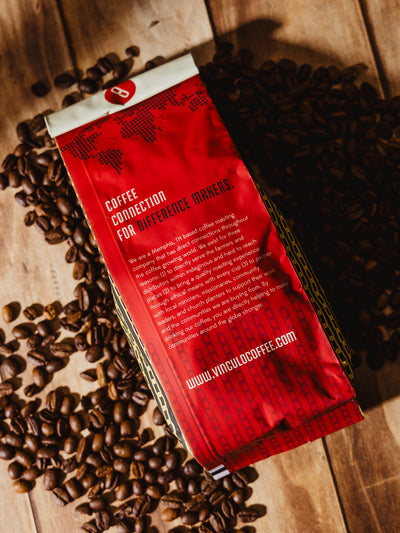 The back of a Vinculo Coffee Roasters bag featuring a bold red design with white text reading "Coffee Connection for Difference Makers." The description highlights the company's mission, ethical sourcing, and community support initiatives. Coffee beans are spread across the wooden surface.