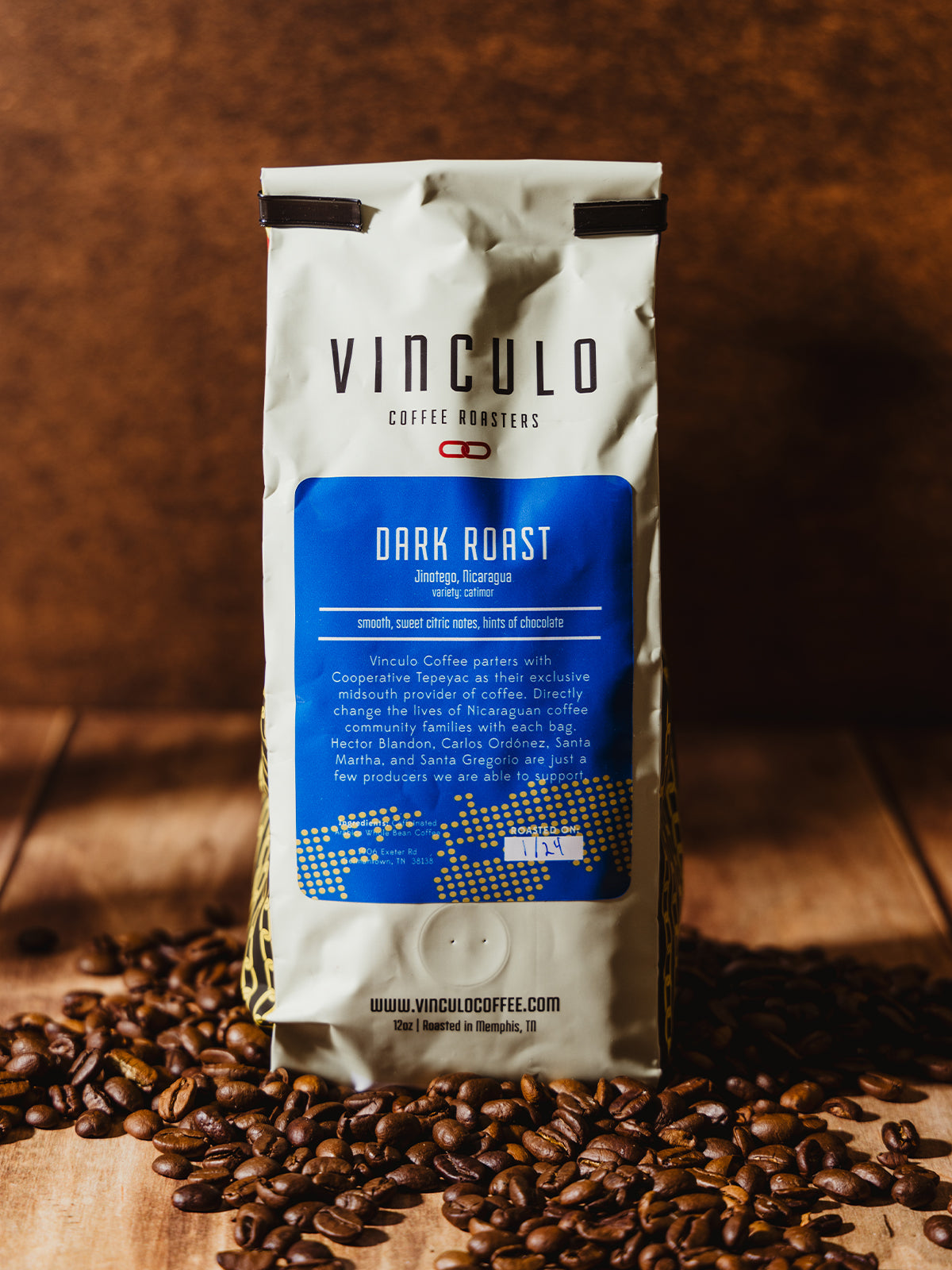 A Vinculo Coffee Roasters Dark Roast coffee bag, displayed on a wooden surface with roasted coffee beans scattered around. The blue label provides tasting notes, and the warm lighting enhances the rich tones of the coffee.